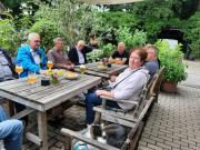 Gruppe von Personen sitzt an einem Holztisch im Freien, umgeben von Pflanzen.